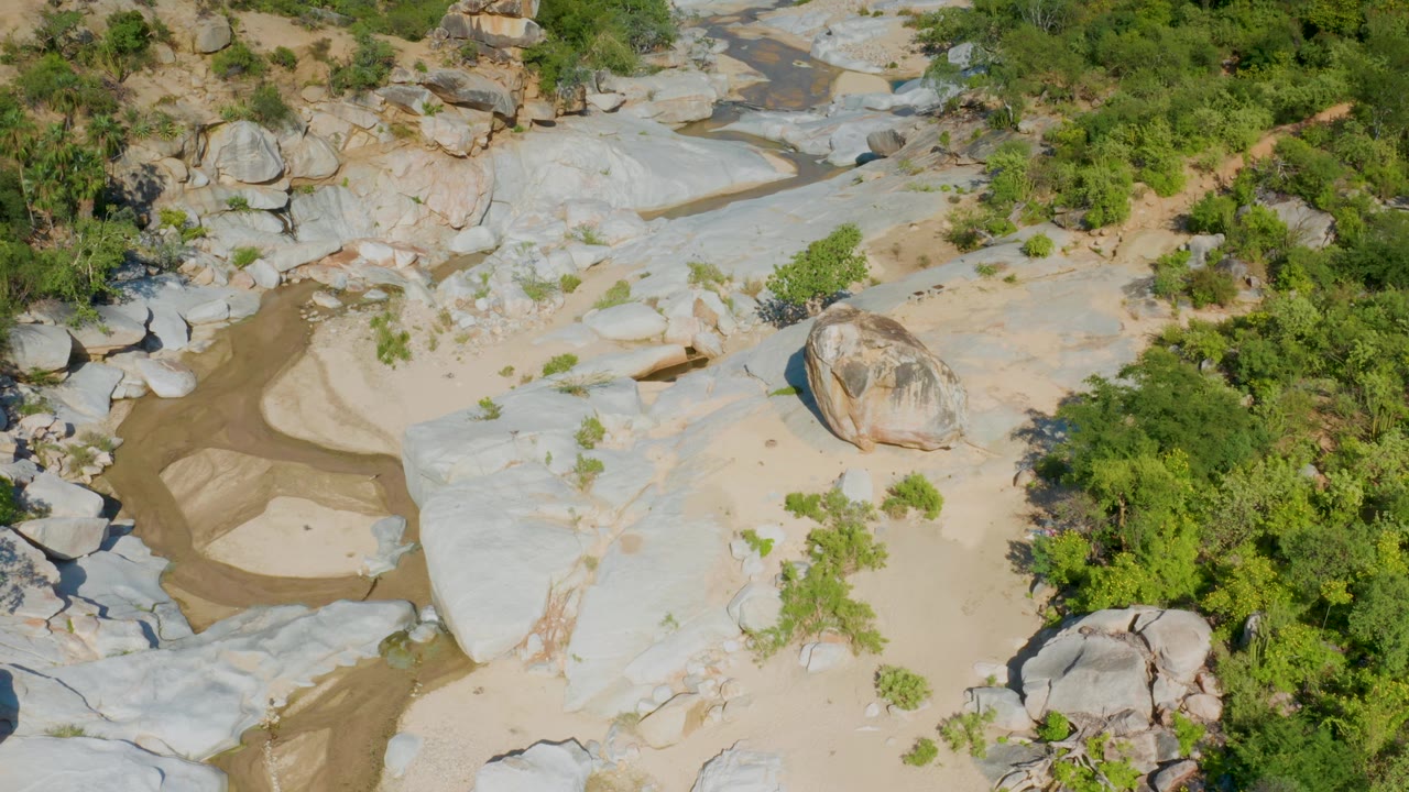Aerial view of a shallow stream beside broad pale rock and dense green brush.