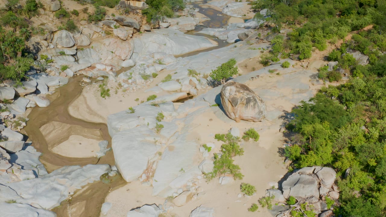 A La Palma aerial frame shows a large rounded boulder near the right-center, pale rock shelves, tan sand pockets, green shrubs, and a thin muddy channel with no visible people.
