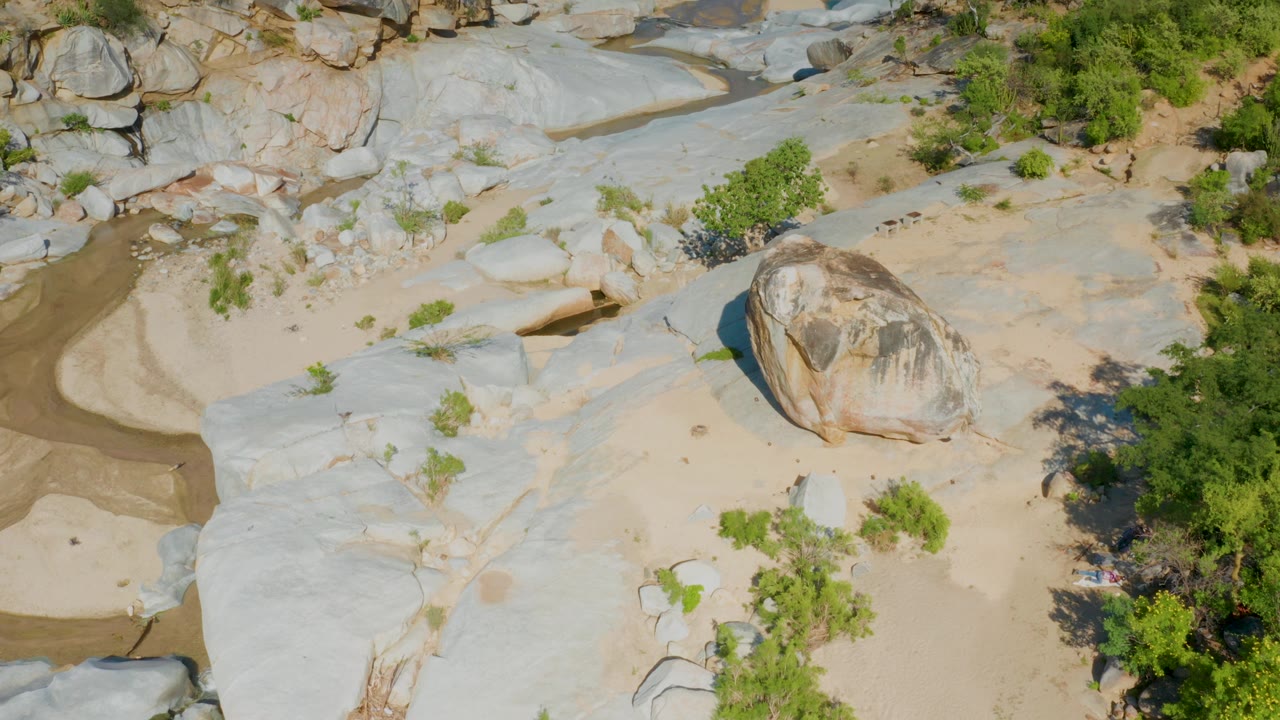 Aerial view in La Palma, Baja California Sur, looking down on a narrow muddy channel cutting through pale rock, tan sand, sparse shrubs, and no visible people.
