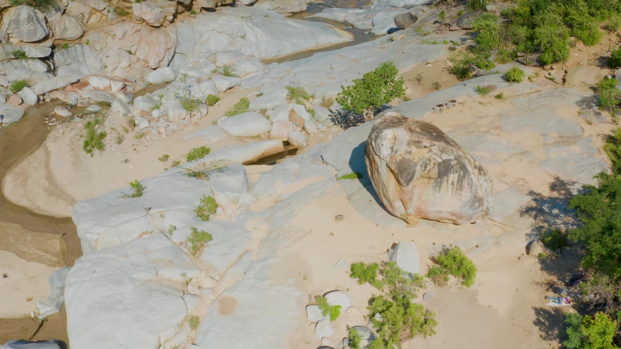 Aerial frame over La Palma, Baja California Sur, with a large boulder set against pale bedrock, tan sand, scattered green shrubs, and no visible people.