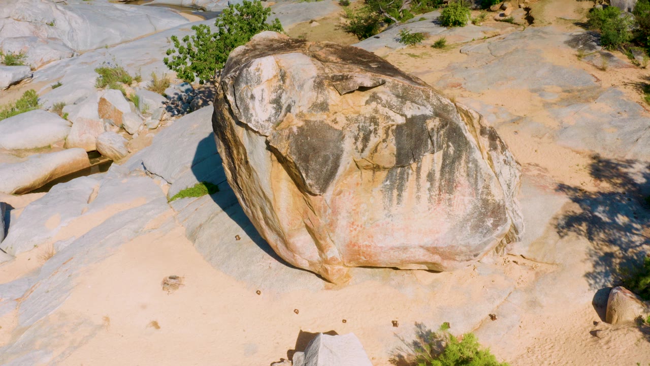 At 0:00, a large tan and gray sandstone boulder sits centered in a sunlit rocky area near La Palma, Baja California Sur, with sandy ground, pale stone slabs, scattered green shrubs, and no visible people.