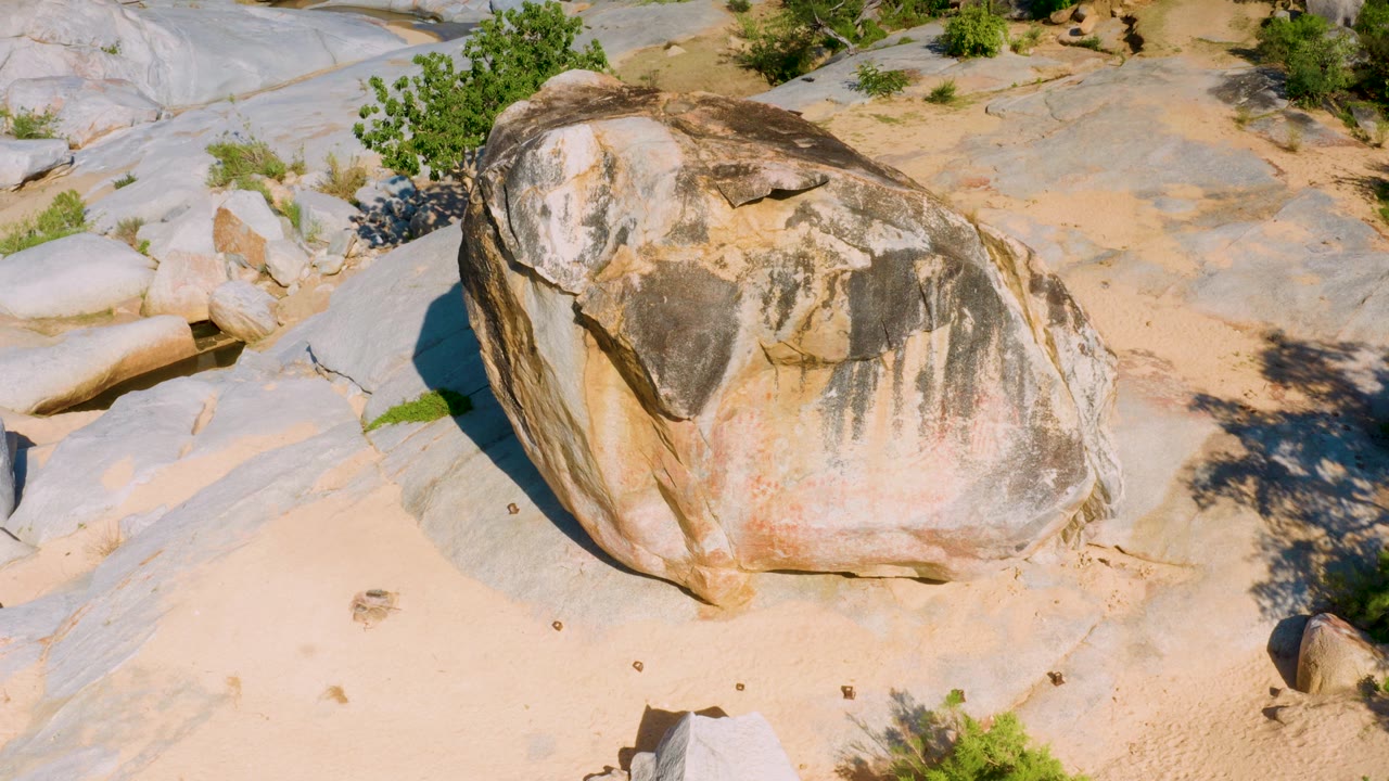 At 0:03, an elevated view keeps the sandstone boulder dominant in the frame, with smooth pale stone shelves, tan sand, a few shrubs, and no visible people in the rocky La Palma setting.