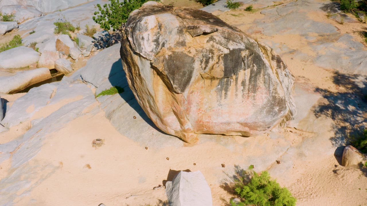 High angle frame of the same brown boulder with darker shadow along its left side, pale stone slabs nearby, green shrubs, and no visible people.