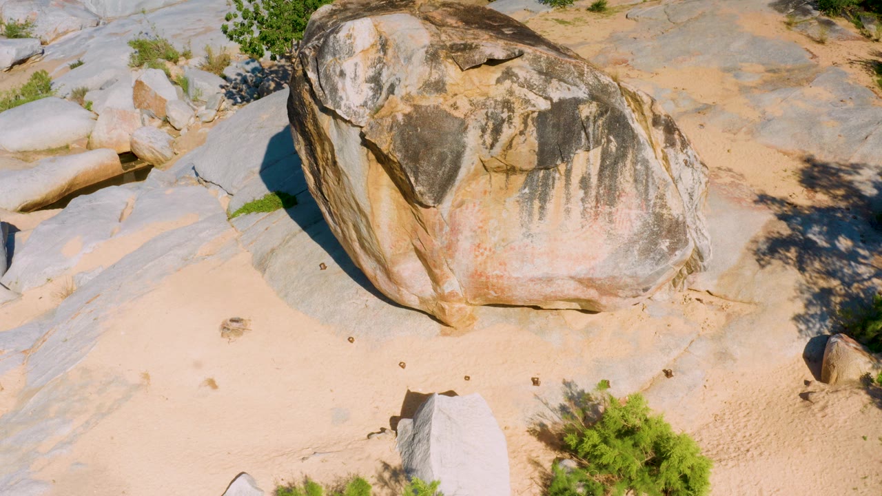 Aerial-looking view of the sunlit boulder on pale desert sand in La Palma, with a small upright rock at right, low green plants, and no visible people.