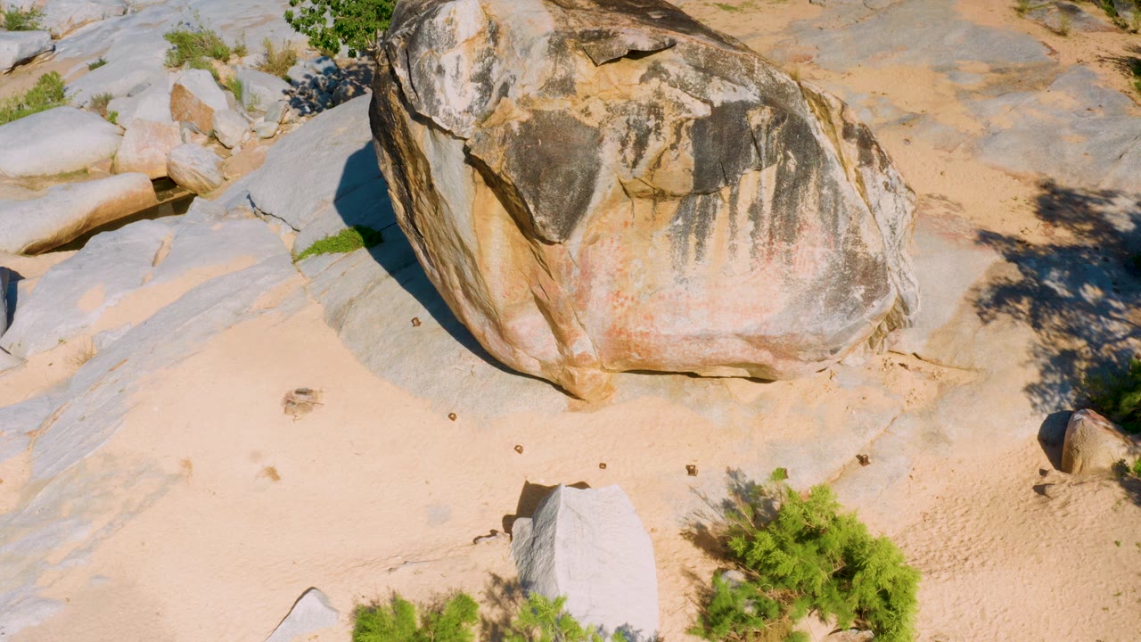 Closer overhead frame of the weathered brown boulder, showing streaked rock faces, pale sand, surrounding stone surfaces, shrubs, and no visible people.