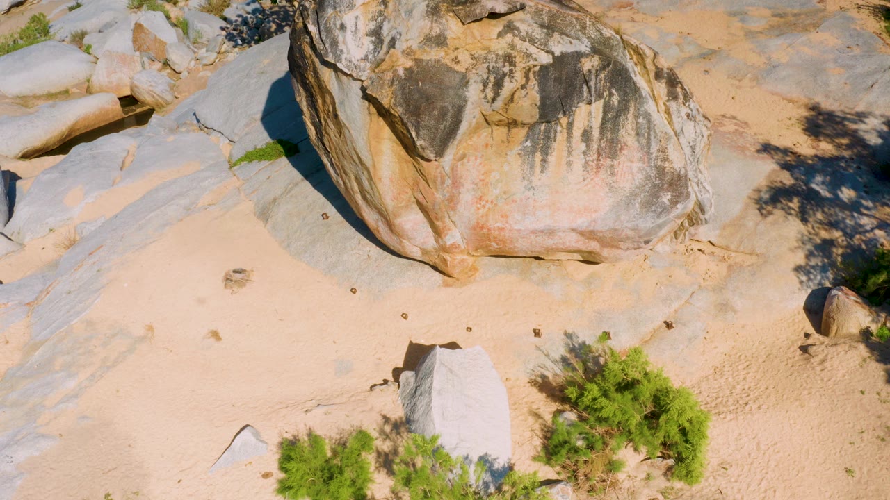 Overhead drone frame in La Palma, Baja California Sur showing a large weathered boulder on pale sand, gray rock slabs at left, green shrubs near the bottom edge, and no visible people.