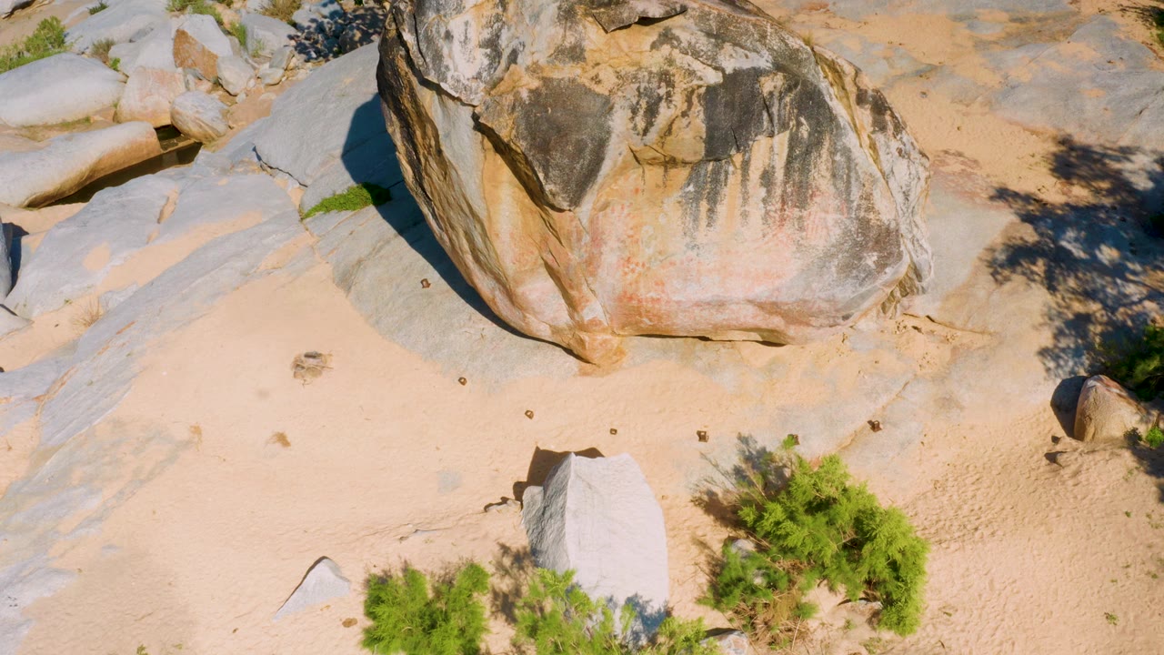 La Palma, Baja California Sur aerial frame with the weathered boulder centered high in frame, pale sand below, a cluster of green shrubs at lower right, and no visible people.