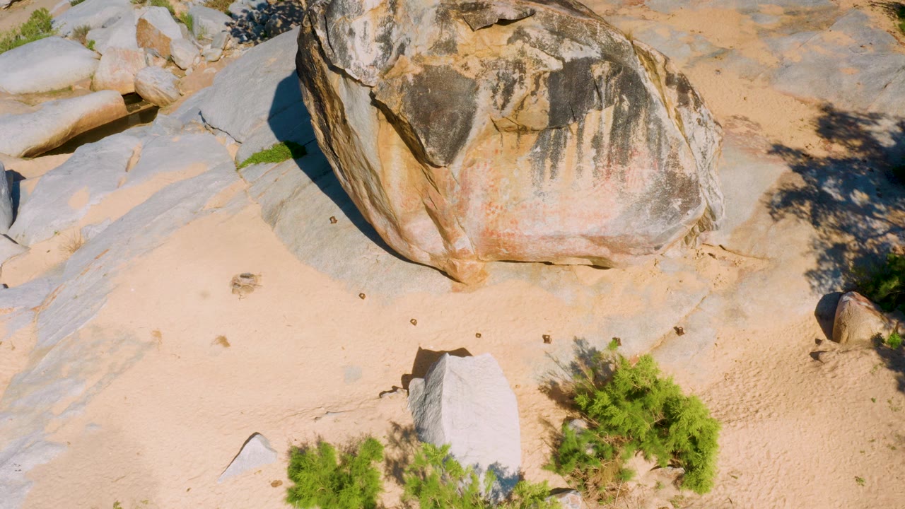 Downward-looking drone frame in La Palma, Baja California Sur with a large tan and gray boulder above pale sand, low shrubs at the lower edge, exposed stone at left, and no visible people.