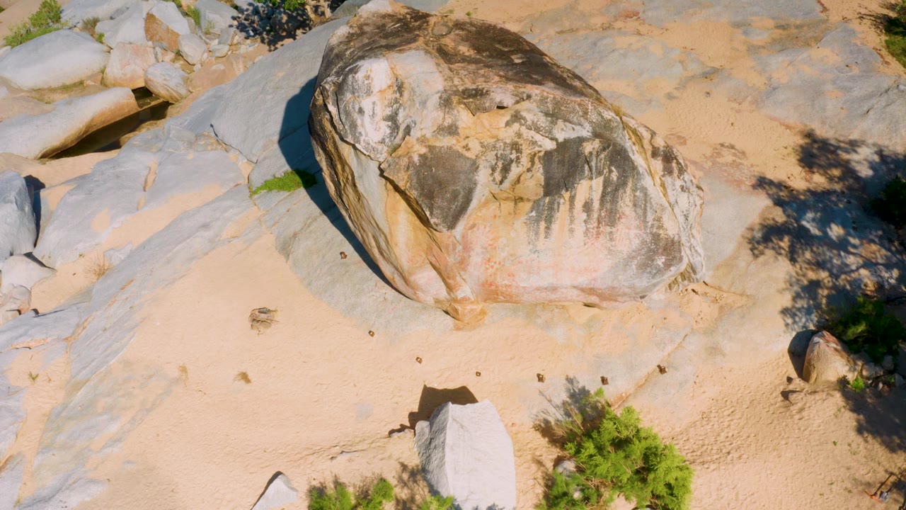 At 0:01 in La Palma, Baja California Sur, the sunlit boulder remains central with pale sand below, smooth gray stone around it, dark shrub shadows to the right, and no visible people.