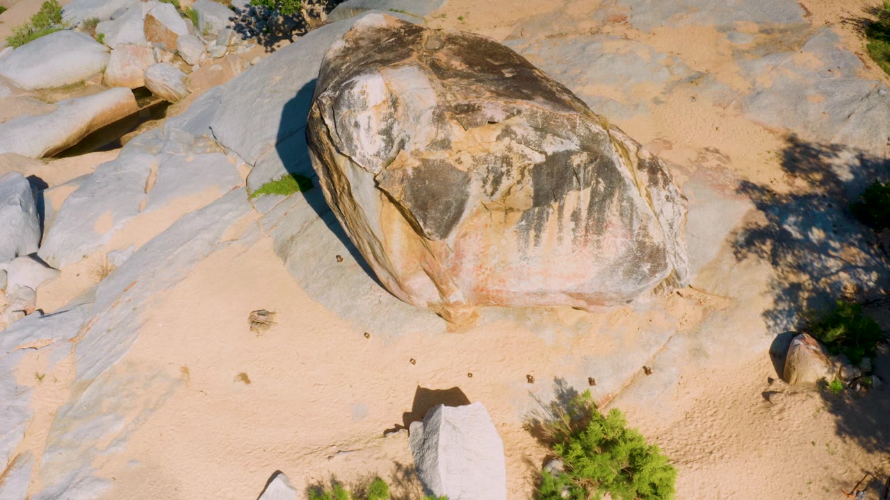At 0:02, the La Palma overhead view shows the boulder slightly right of center, pale sand in the lower half, broad gray rock slabs behind it, small green shrubs, and no visible people.