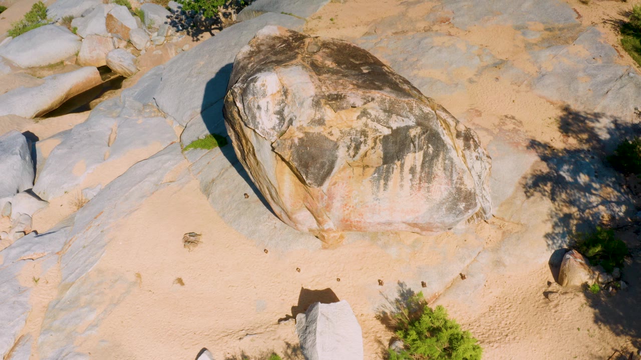 At 0:03 in La Palma, Baja California Sur, the boulder fills the middle of the frame with a dark shaded side, sandy ground below, exposed stone around it, shrub shadows at right, and no visible people.