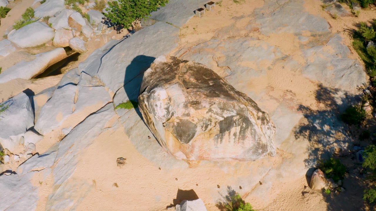 Overhead frame in La Palma, Baja California Sur showing a large weathered boulder on sand, gray rock slabs, green shrubs near the edges, hard sunlight, and no visible people.