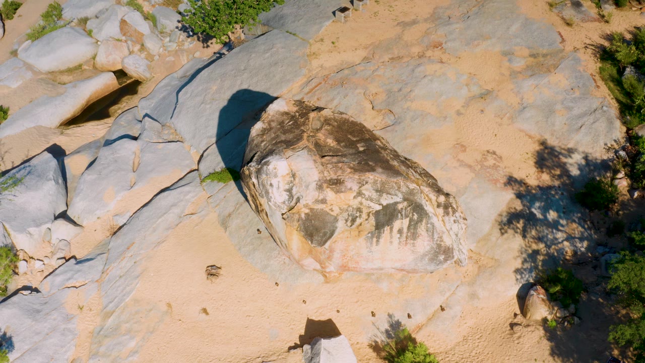 Top-down frame in La Palma, Baja California Sur showing the central stone slightly lower in the composition, sandy terrain, large gray slabs, green bushes to the right, and no visible people.