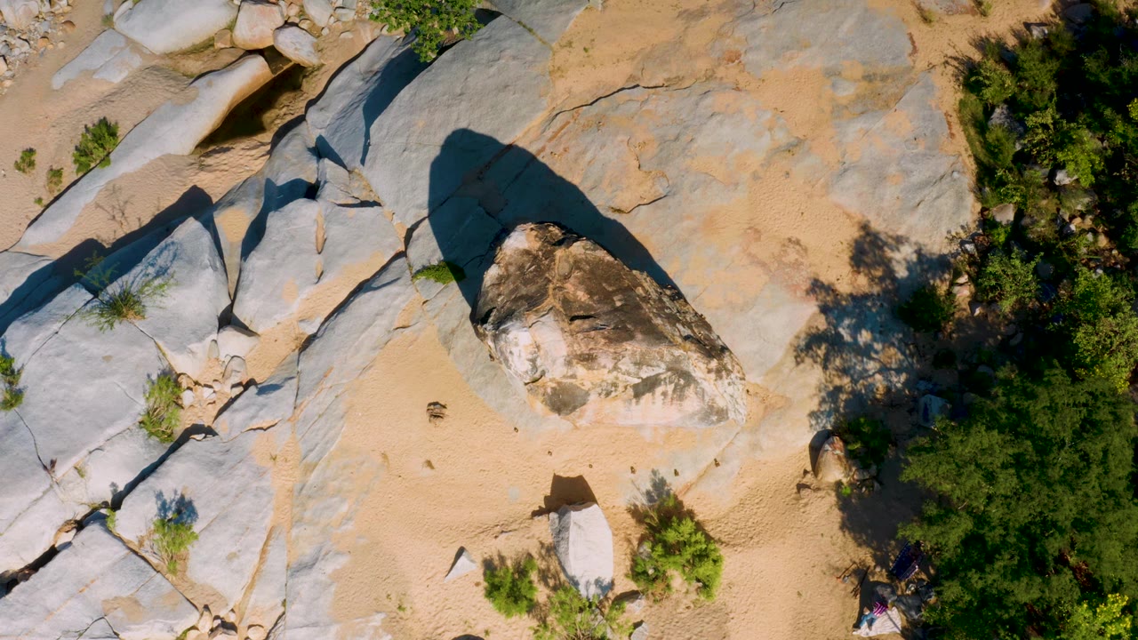 Aerial frame in La Palma, Baja California Sur with a rounded central boulder, pale stone sheets, tan sand pockets, shrubs, and no visible people.