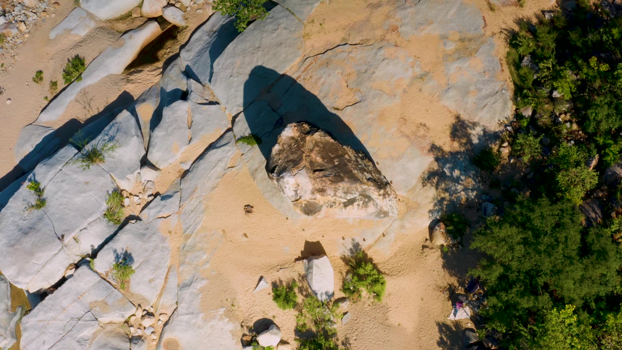Overhead frame from La Palma, Baja California Sur with pale stone planes on the left, a shadowed boulder near center, tan sand below, green vegetation to the right, and no visible people.