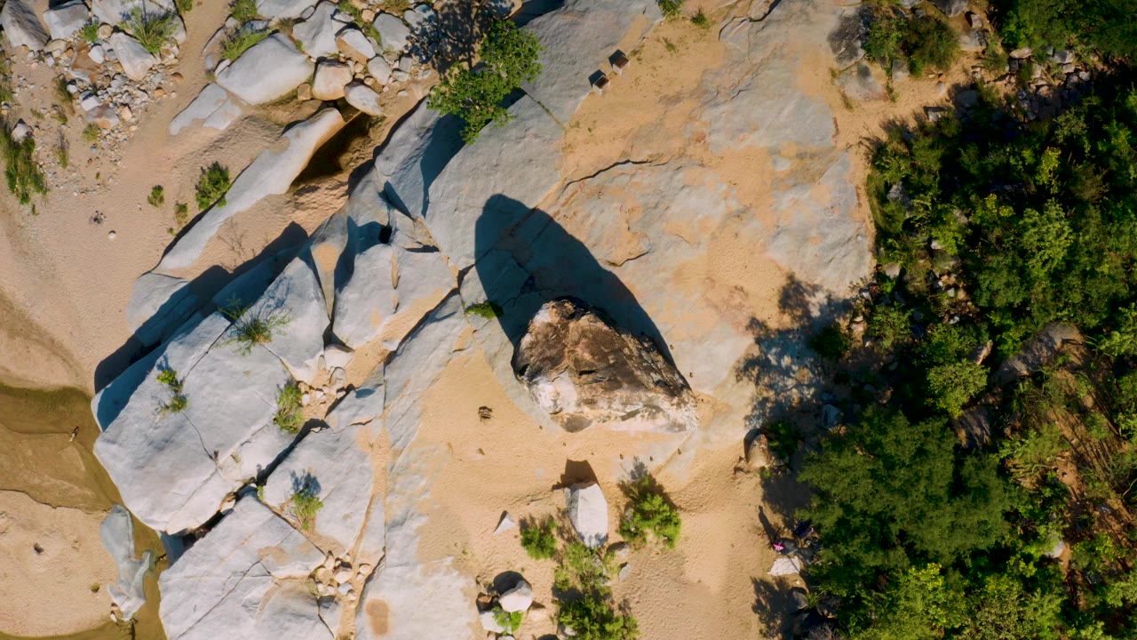 Overhead aerial frame from La Palma, Baja California Sur, showing pale rock slabs beside sandy ground, dense green vegetation, a narrow wash, and no visible people.