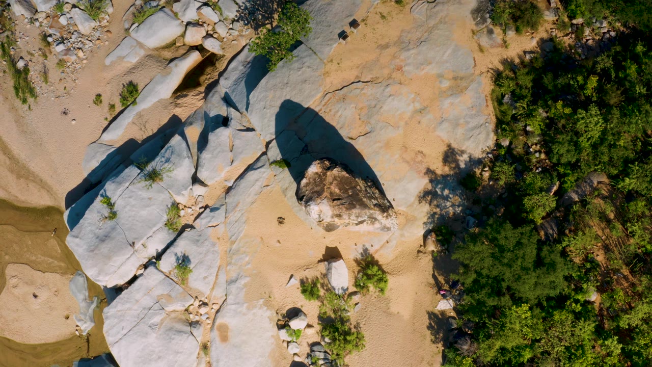 Top-down frame in La Palma, Baja California Sur, showing a large shadowed boulder on pale stone, sandy open ground, green vegetation, and no visible people.
