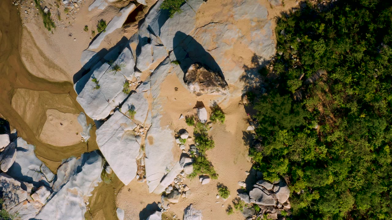 Aerial frame over La Palma terrain with white-gray rock slabs, beige sand patches, small stones, dense green trees, and no visible people.
