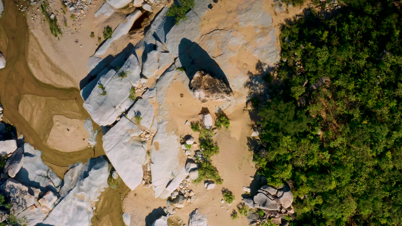 Overhead La Palma frame showing a curved shallow creek area at left, pale boulders and sandy ground in the center, thick trees to the right, and no visible people.