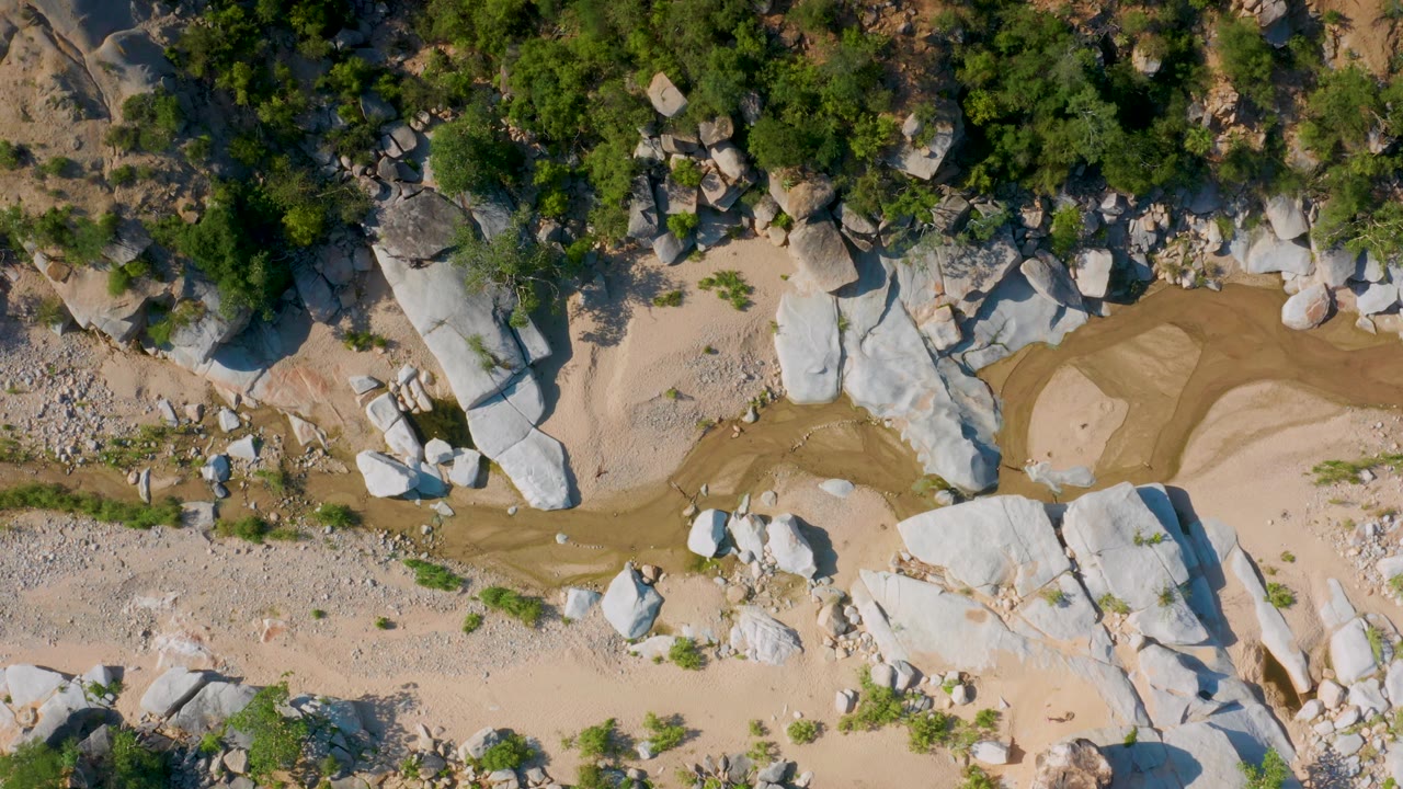 Top-down La Palma frame showing clustered white rocks, patchy green vegetation, beige sand, a narrow tan wash, and tiny visible people on the ground.