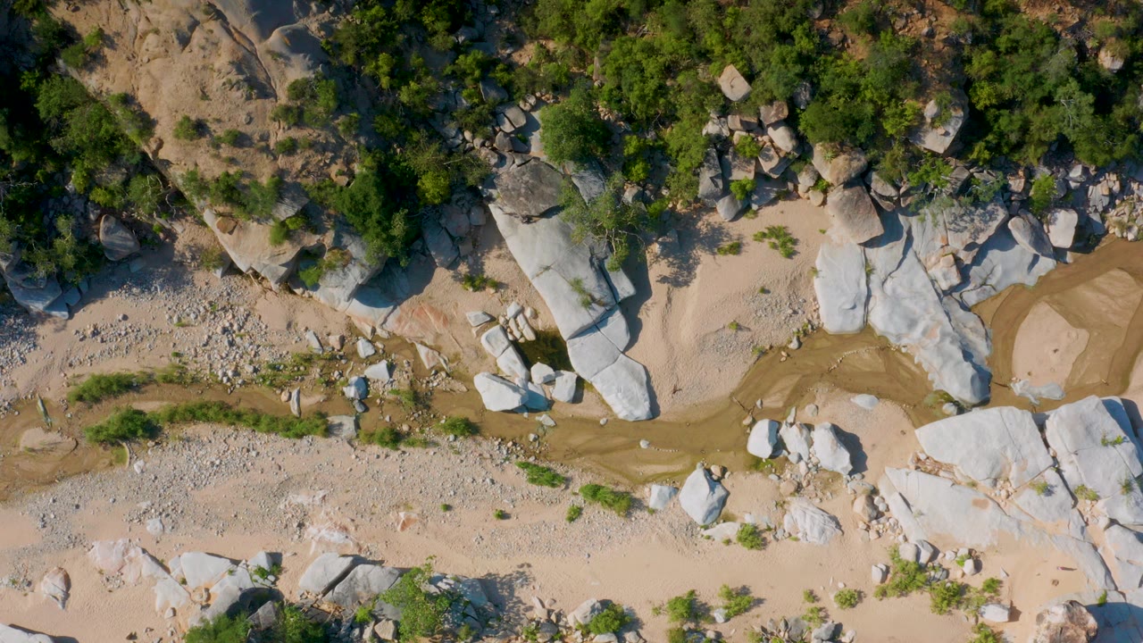 Overhead view in La Palma, Baja California Sur, showing tan sand, pale rock slabs, green shrubs, scattered stones, and a narrow muddy channel with no visible people.