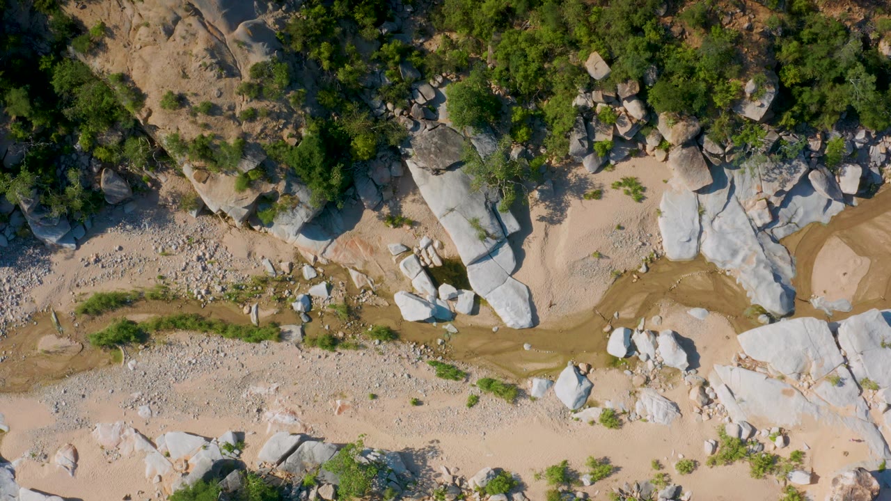 Top-down view in La Palma, Baja California Sur, where shrubs line the upper slope and a winding sandy channel passes between boulders, with no visible people.