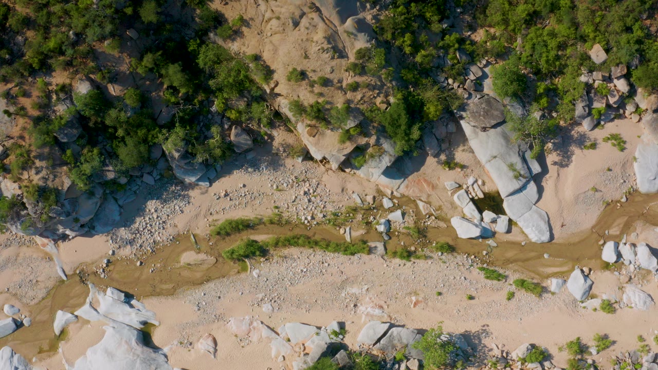 Overhead frame from La Palma, Baja California Sur showing pale sand, a narrow ravine, scattered stones, large boulders, green trees, and no visible people.