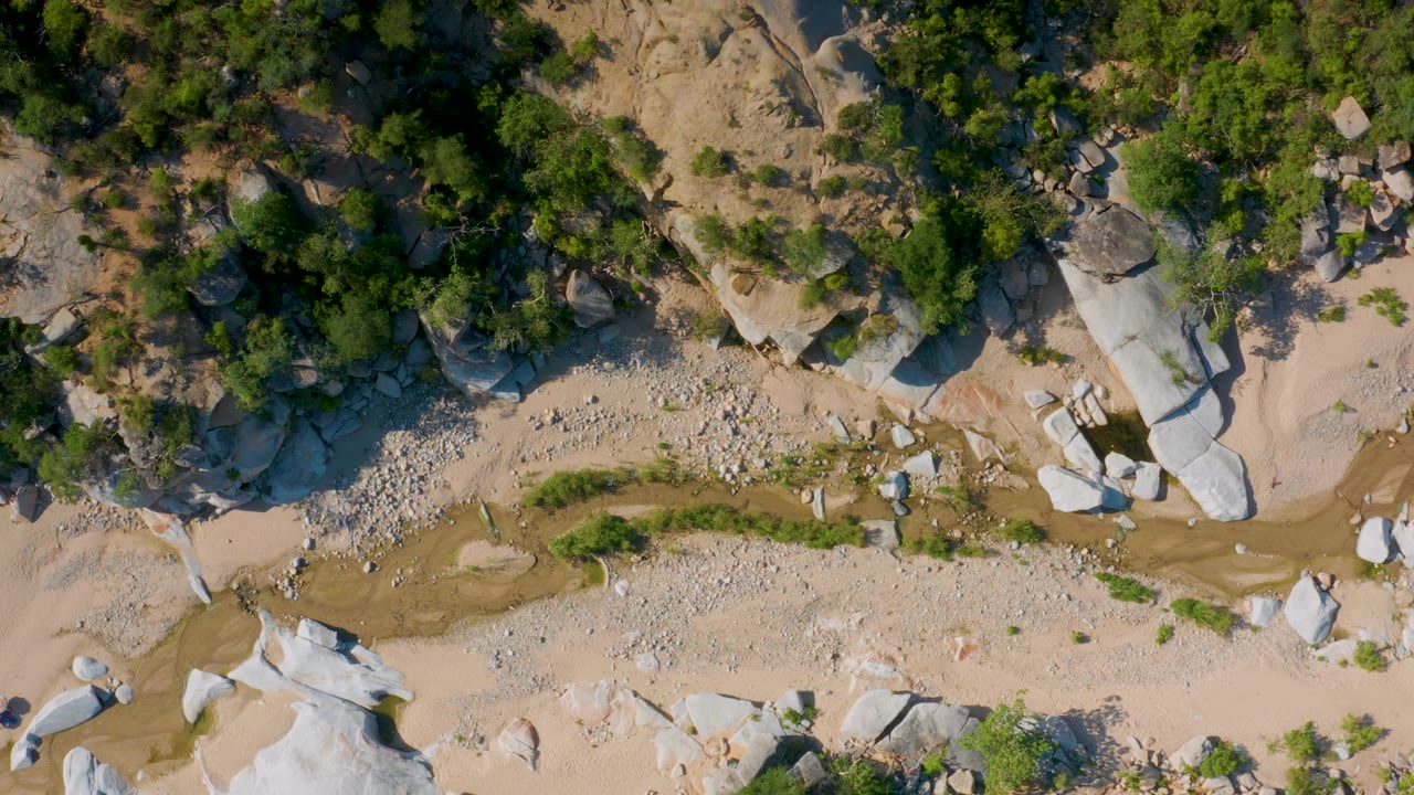 Aerial view in La Palma, Baja California Sur with a tan ravine, clusters of small stones, rounded pale boulders, green vegetation, and no visible people.