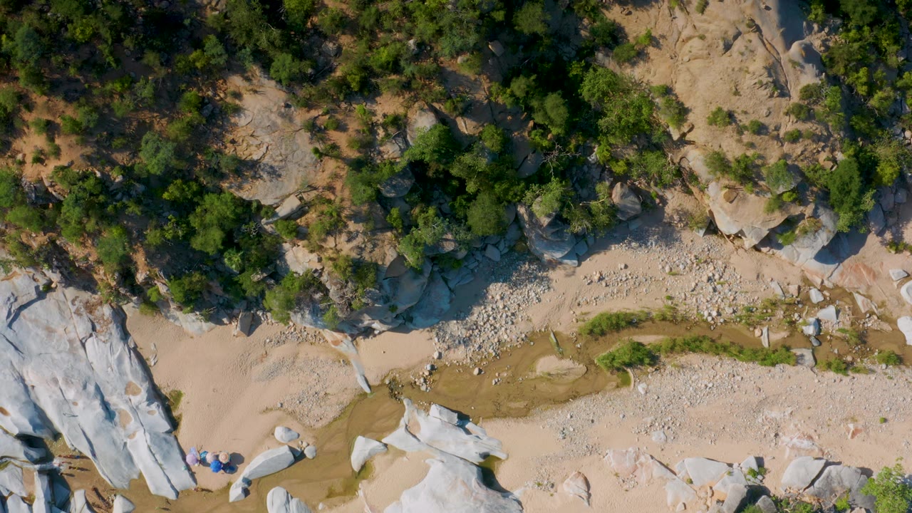 Aerial frame over La Palma, Baja California Sur showing white rock outcrops on the lower right, a tan wash through the middle, green trees above, and no visible people.