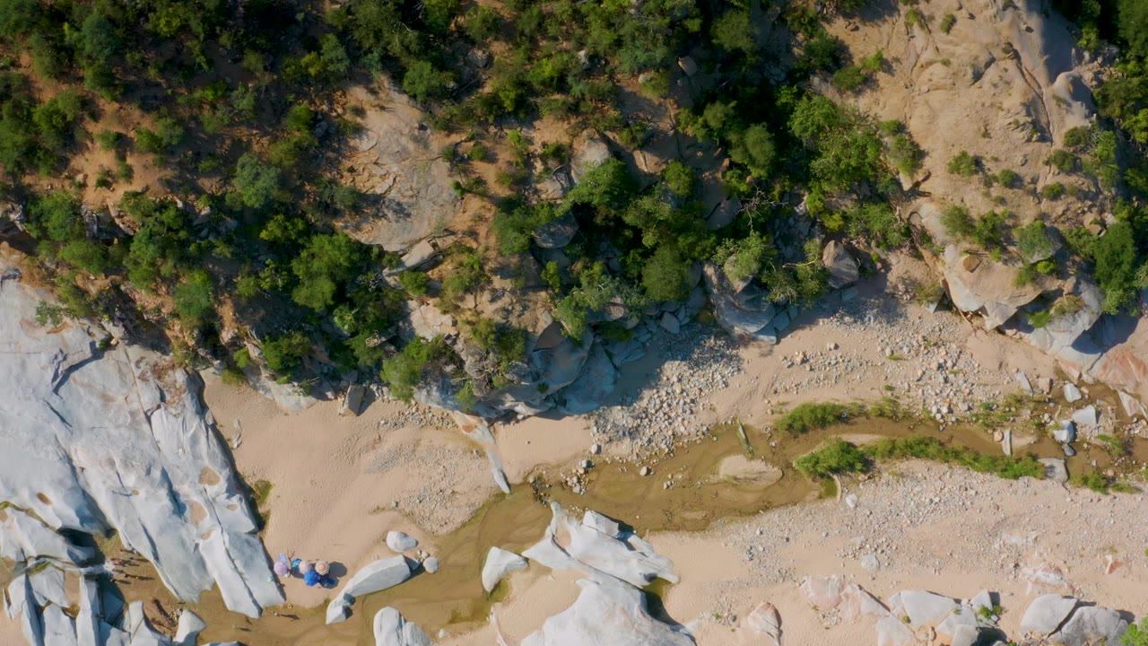 Overhead La Palma, Baja California Sur frame with a large pale rock slab along the left edge, sandy ground below, gravel patches, green shrubs, and no visible people.