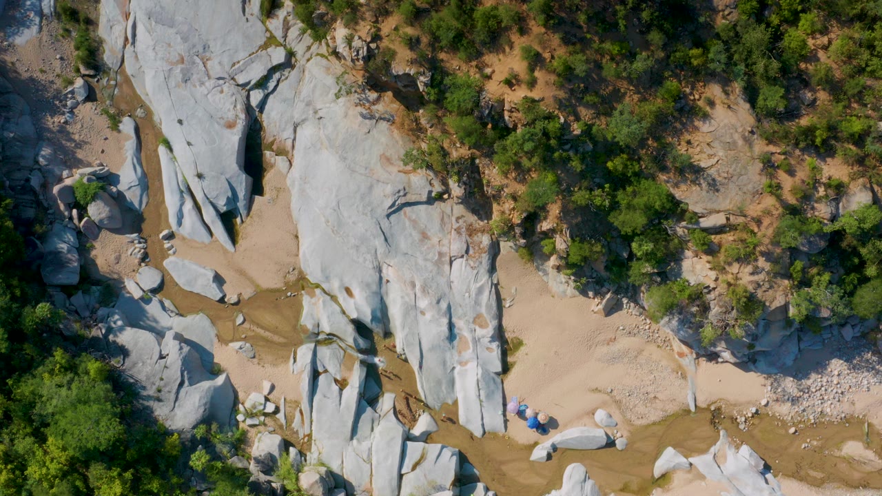 Overhead frame from La Palma, Baja California Sur showing pale vertical rock slabs, sandy wash areas, green shrubs, and shallow brown water channels, with no visible people.