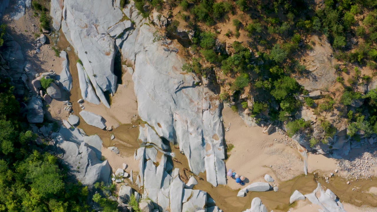 Top-down La Palma, Baja California Sur terrain with a broad pale stone formation, sandy pockets, small boulders, and a darker forest edge, with no visible people.