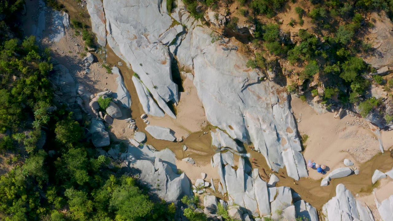 Top-down frame over La Palma, Baja California Sur, showing pale stone ridges, tan sand, scattered boulders, small brown-green pools, surrounding trees, and no visible people.