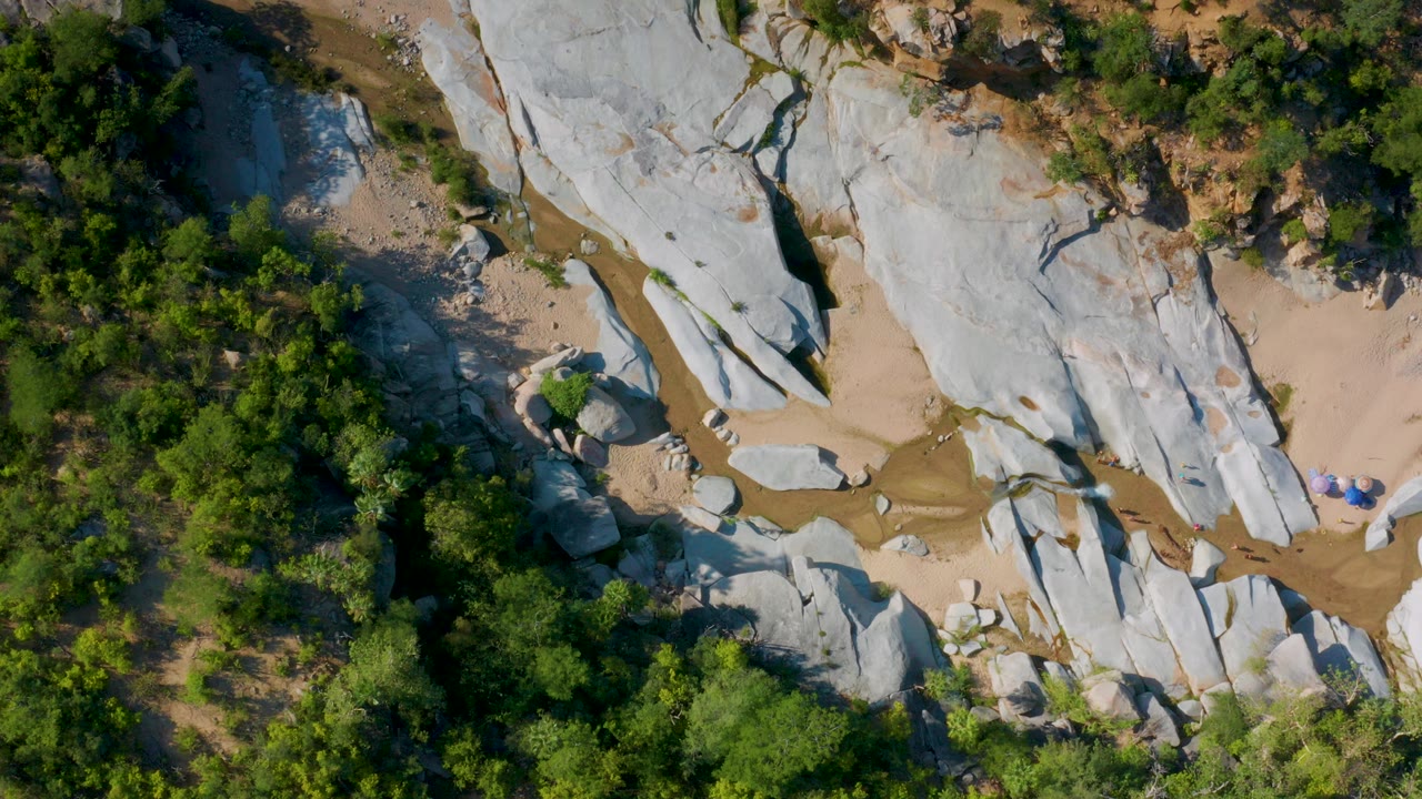 Top-down aerial frame in La Palma, Baja California Sur showing pale rock slabs, tan sand, a narrow muddy channel, green shrubs, and tiny distant people near the right side.