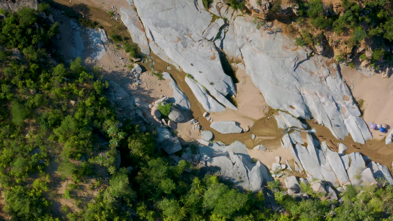 Top-down frame in La Palma, Baja California Sur showing smooth pale stone, scattered boulders, tan sand, a thin watercourse, dense greenery, and very small visible people on the right.