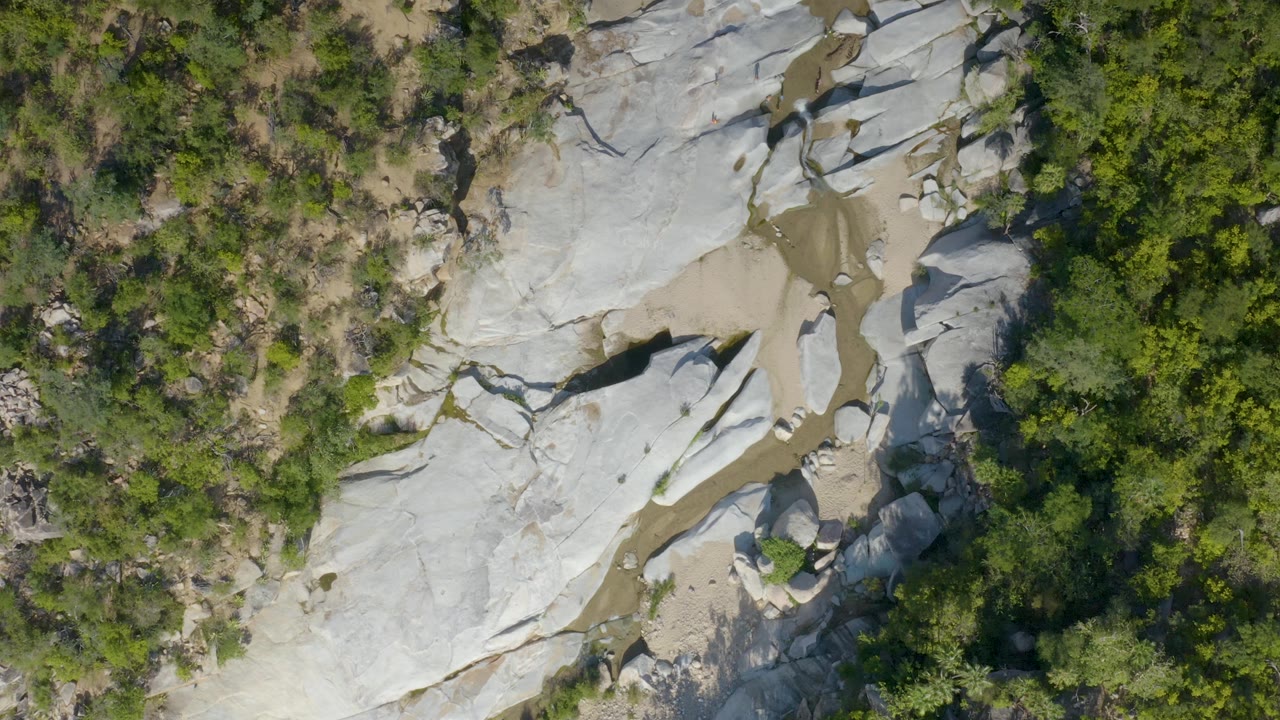 Aerial frame in La Palma, Baja California Sur with pale stone shelves on the left, a brown muddy channel through the middle, and dense green vegetation at the edge with no visible people.