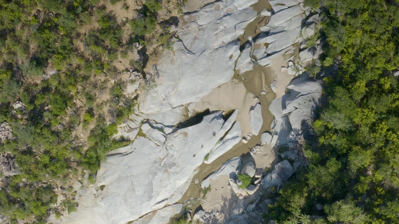 Top-down La Palma, Baja California Sur view showing a broad white rock formation, small muddy pools or wet sand, and thick green brush with no visible people.