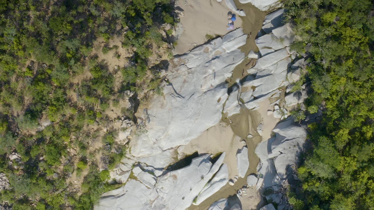 Overhead frame from La Palma, Baja California Sur showing smooth pale rock slabs, tan sand, and dense green brush along a narrow ravine, with no visible people.