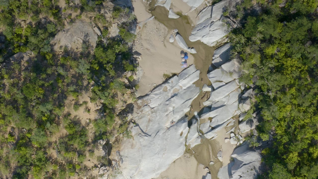 High overhead view in La Palma, Baja California Sur with a muddy creek line running between light-colored rock formations and green brush, with no visible people.
