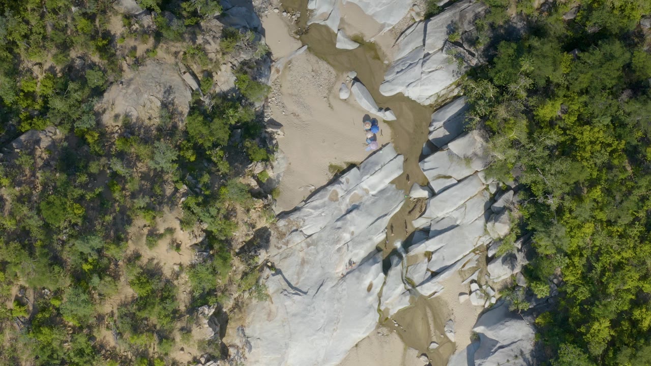 Top-down frame over La Palma terrain showing pale rock shelves, a muddy water trace, tan sand, and dense shrubs around the wash, with no visible people.