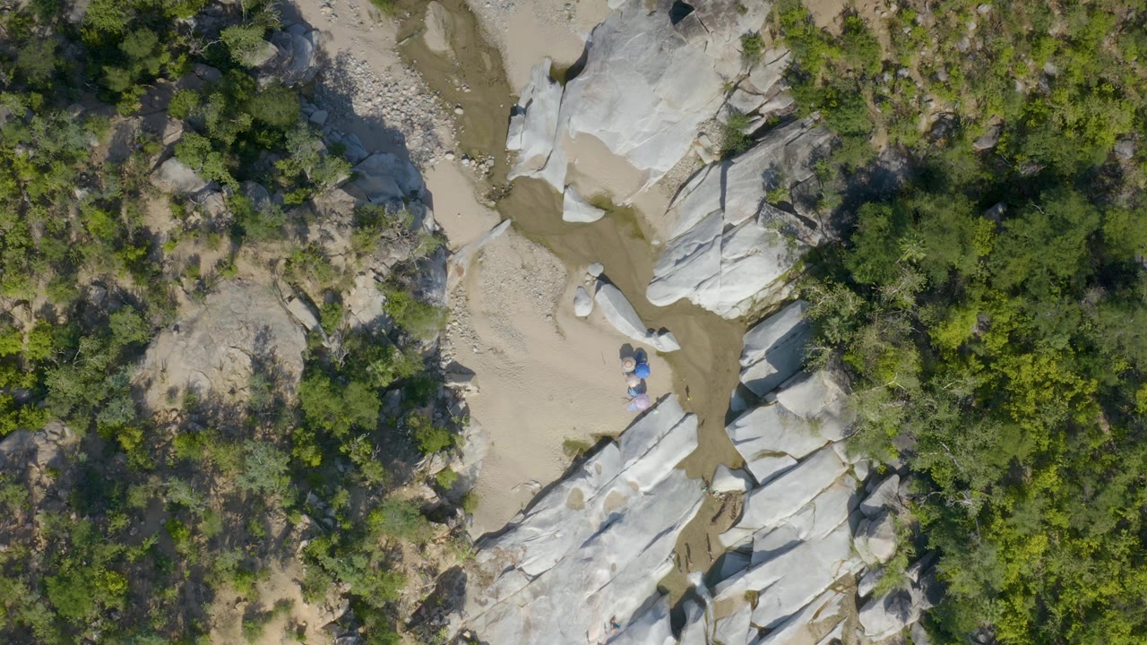 Overhead La Palma, Baja California Sur view with scrubland on one side and pale rock bordering a sandy wash with small muddy water sections, with no visible people.