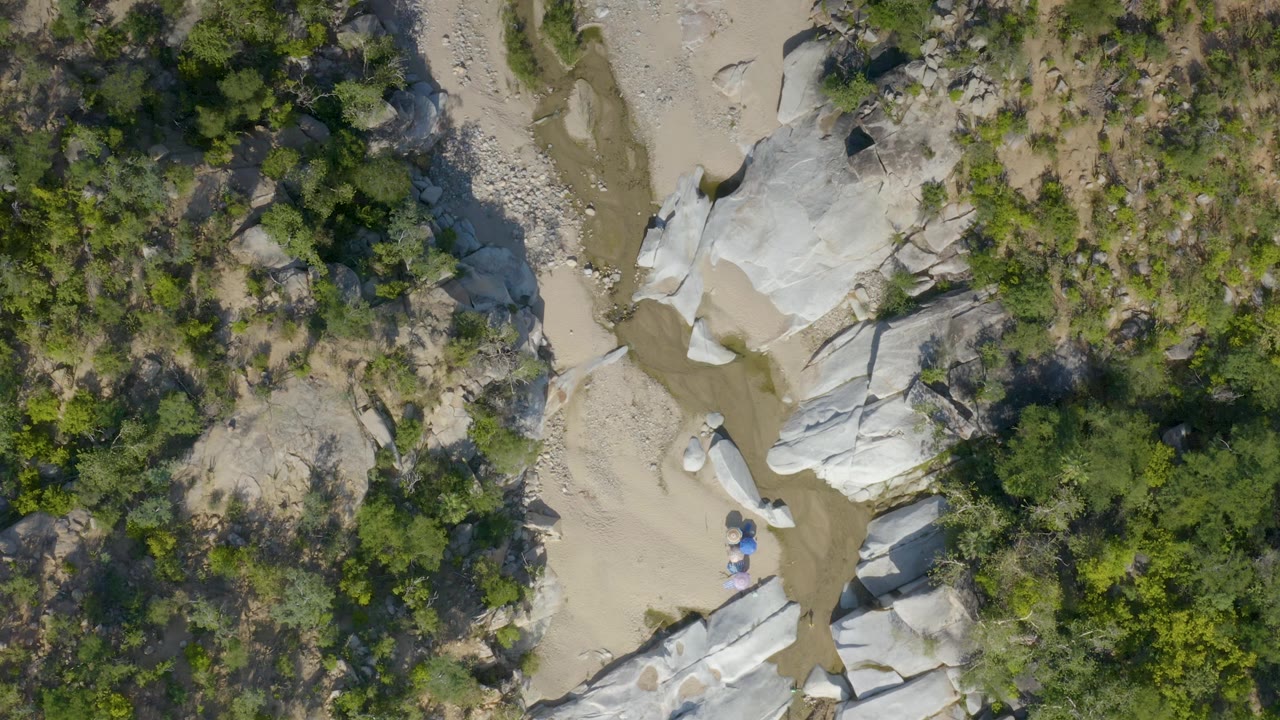 Aerial frame over La Palma, Baja California Sur with pale stone outcrops close to a tan water channel and scrubby green forest filling the surrounding ground, with no visible people.