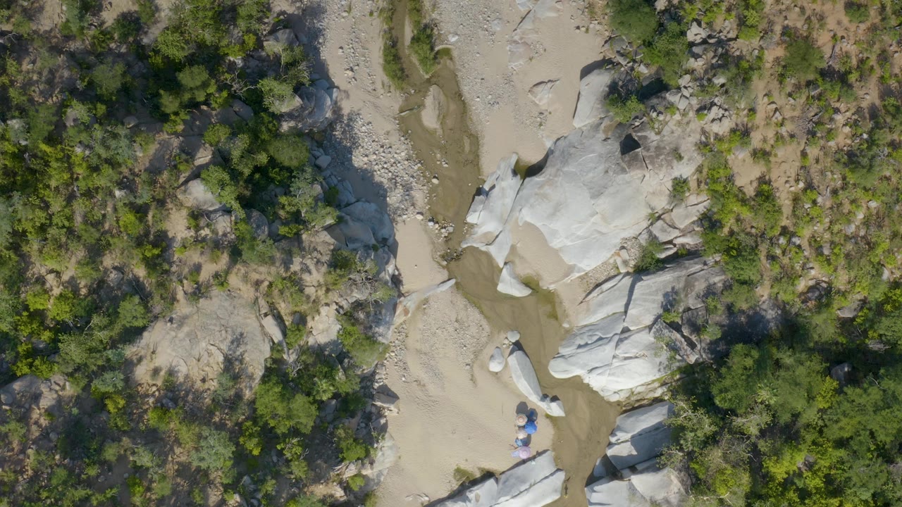 Aerial still from La Palma, Baja California Sur where a narrow dry creek channel curves past smooth pale rocks and clustered trees, with no visible people.