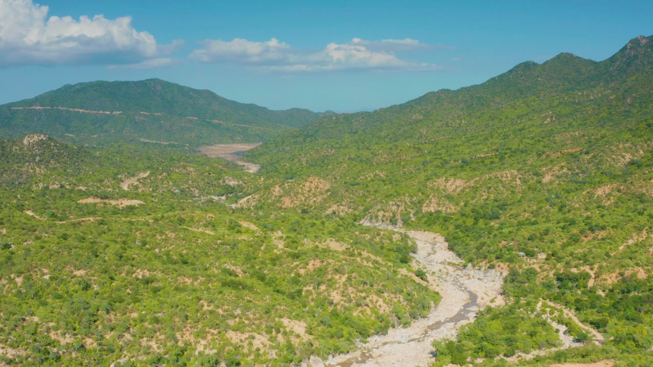 High aerial view over La Palma, Baja California Sur, with green scrubland, rocky hills, a pale dry wash in the foreground, distant mountain ridges, blue sky, and no visible people.