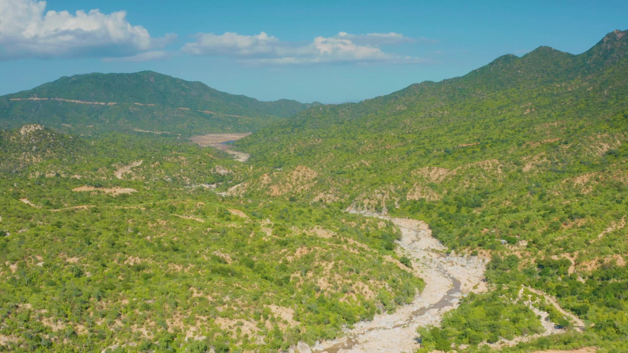 Wide overhead view of La Palma, Baja California Sur, with a dry channel crossing the lower frame, scrub-covered slopes in the middle distance, mountains at the horizon, and no visible people.