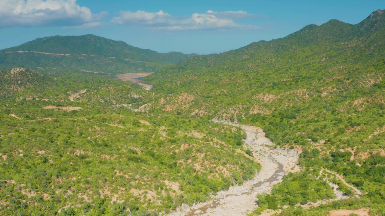 Sunlit aerial view in La Palma, Baja California Sur, looking across green arid hills, a pale riverbed or dirt track in the lower frame, mountain ridges, and no visible people.