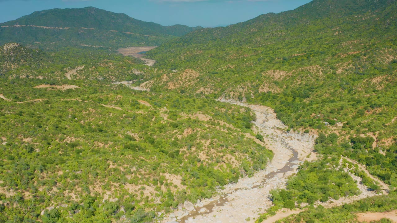 Aerial view in La Palma, Baja California Sur, with a pale dry riverbed running from the foreground into green slopes and layered mountain ridges, with no visible people.