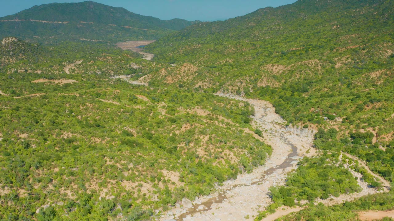 Aerial frame from La Palma, Baja California Sur, showing the winding light-colored wash between brush-covered hills, with darker mountains along the horizon and no visible people.