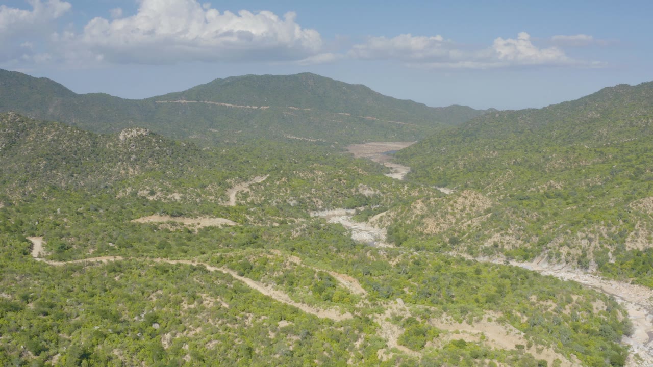 Aerial view near La Palma, Baja California Sur, showing green hills, pale dirt roads, rocky drainage lines, distant mountains, blue sky, and no visible people.