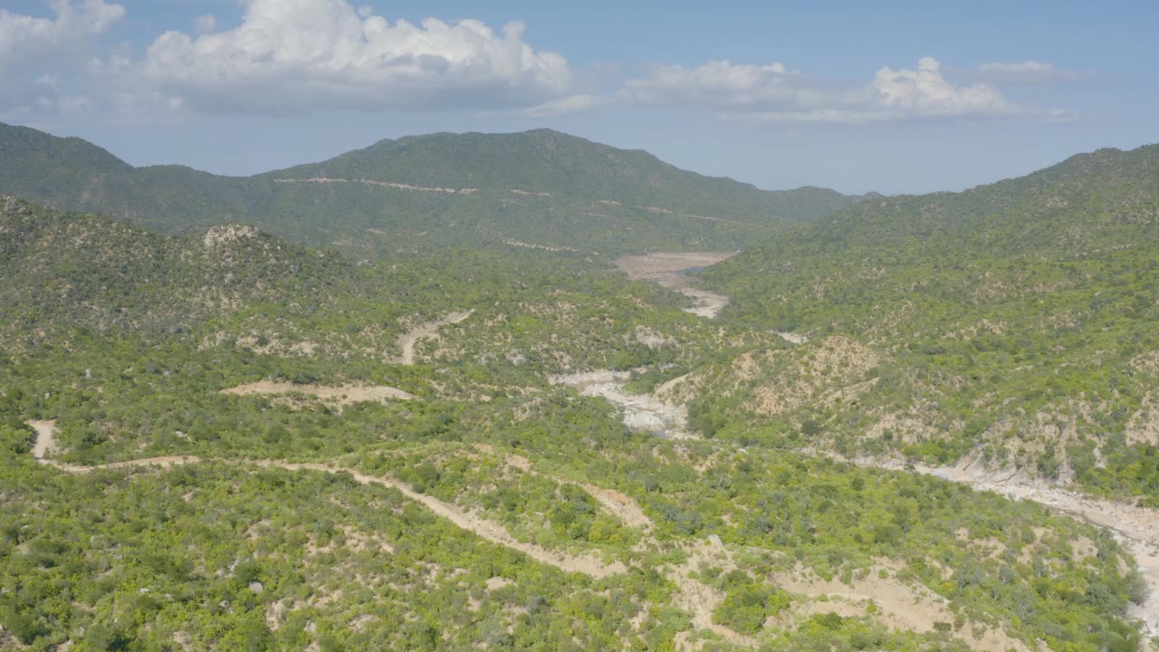 Aerial frame near La Palma, Baja California Sur, showing green hills in the foreground, pale tracks and drainage cuts in the midground, a broad ridge beneath a large cloud, and no visible people.